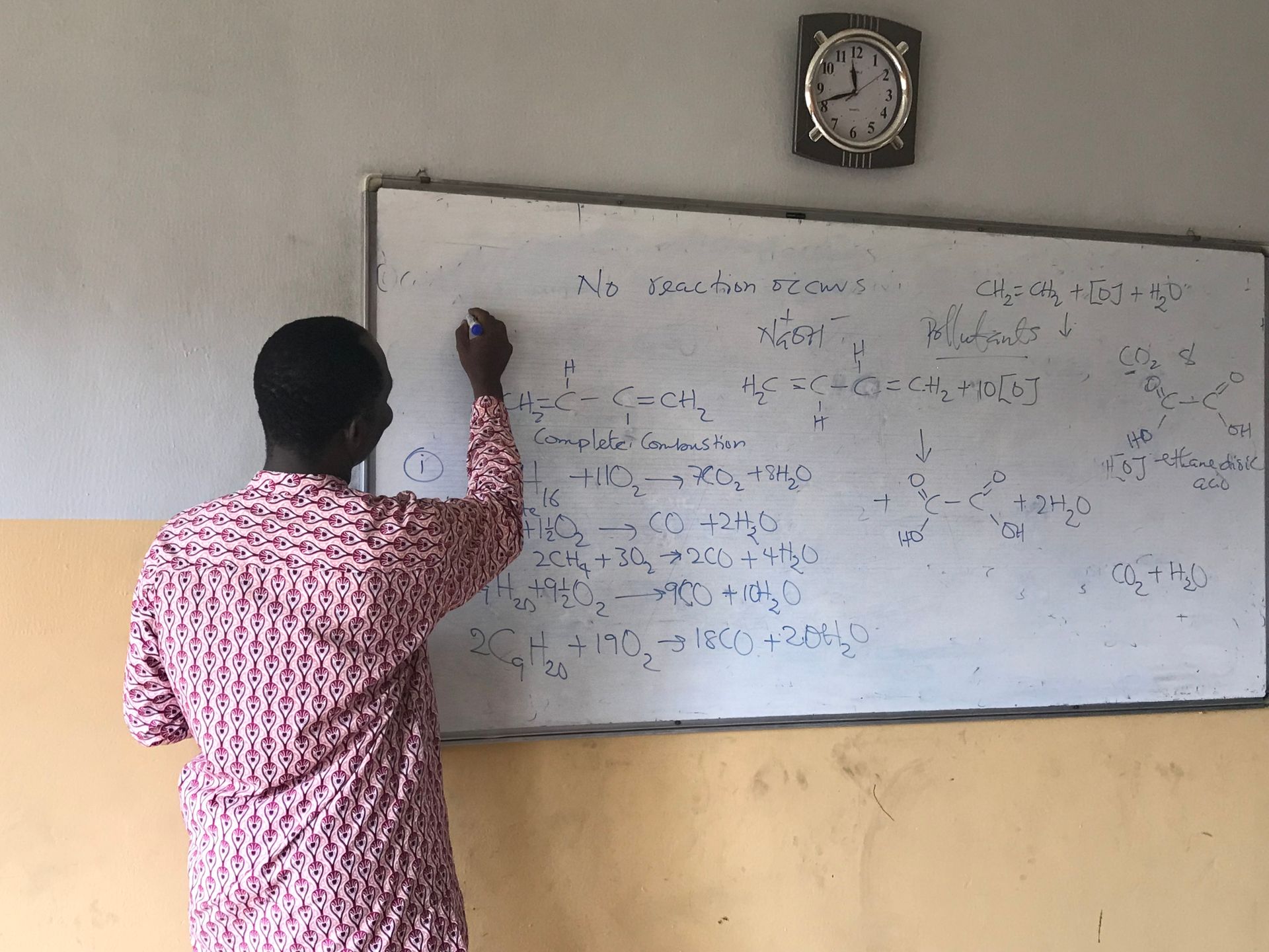 Male teacher writing chemical equations on a classroom whiteboard.