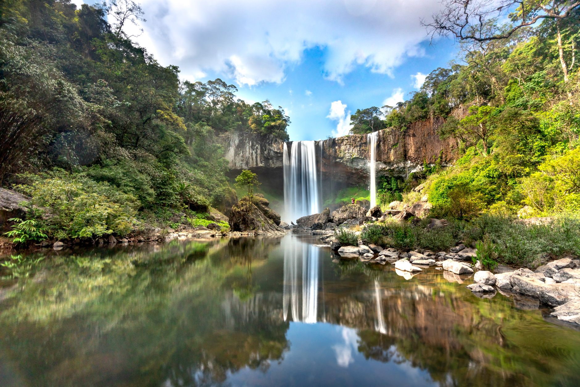 Stunning waterfall with serene reflections surrounded by lush greenery under a blue sky.