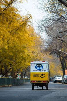 An urban autumn scene in Beijing featuring a delivery vehicle on a tree-lined street with golden foliage.