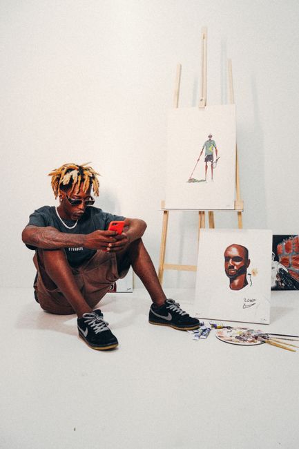 A young artist in Brazil sits on the floor, browsing his phone, surrounded by his paintings and art supplies.
