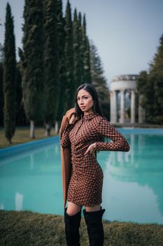 Stylish woman in patterned dress poses confidently near a serene pool surrounded by trees.
