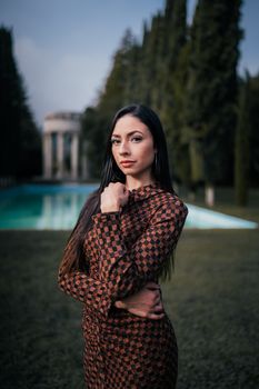 A woman in a patterned dress poses gracefully by a pool with trees and a building in the background.