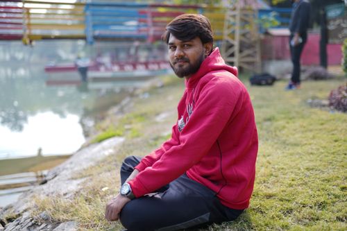 Free Portrait of a man in a red hoodie sitting on grass by a serene pond. Stock Photo