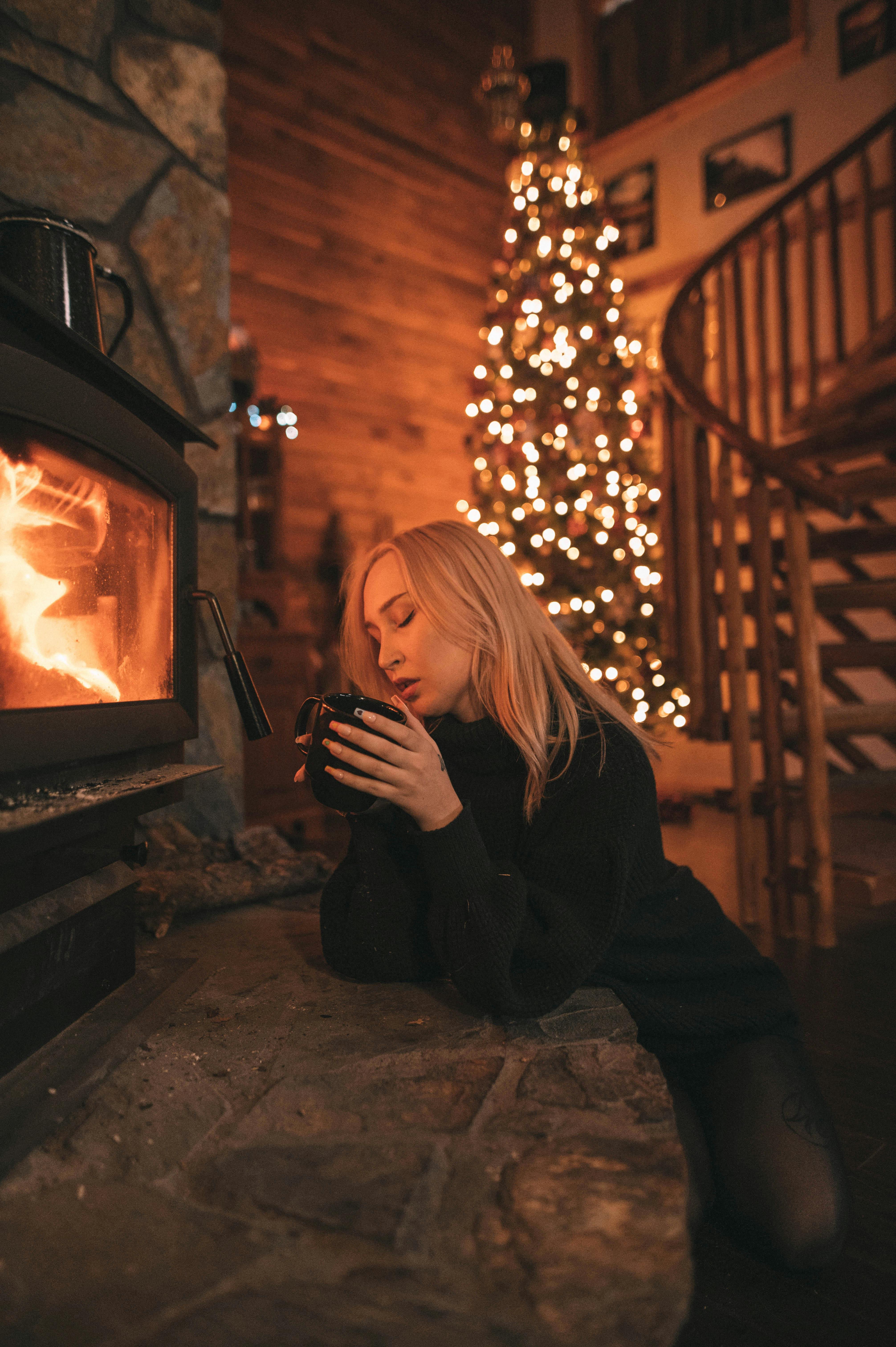 A woman enjoys a warm drink by the fireplace beside a Christmas tree, embodying winter coziness.