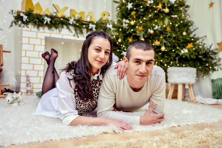 Couple Lying On White Carpet In Front Of Fireplace And Christmas Tree