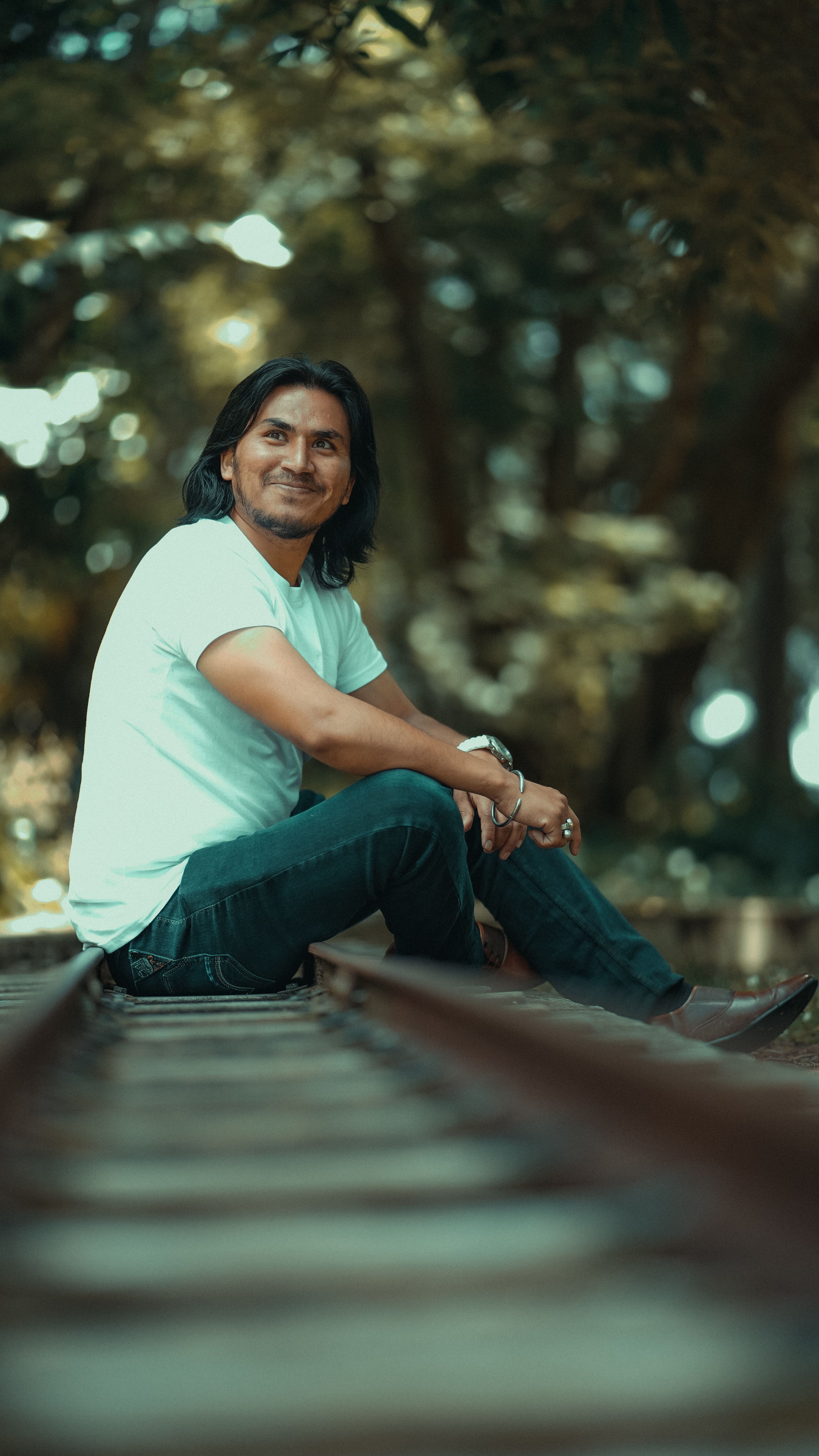 Smiling man with long hair sitting on railroad tracks surrounded by nature.
