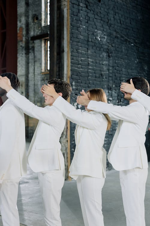 Group of People in White Clothes Standing in a Line and Covering Eyes of the Person in front of Them 