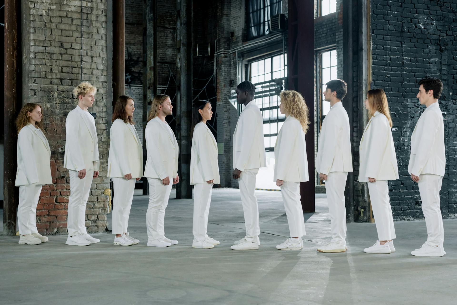 A diverse group of adults dressed in white suits standing in an industrial warehouse setting, facing each other.