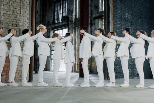 Group of People in White Clothes Standing in a Line and Covering Eyes of the Person in front of Them 