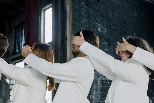Group of People in White Clothes Standing in a Line and Covering Eyes of the Person in front of Them 