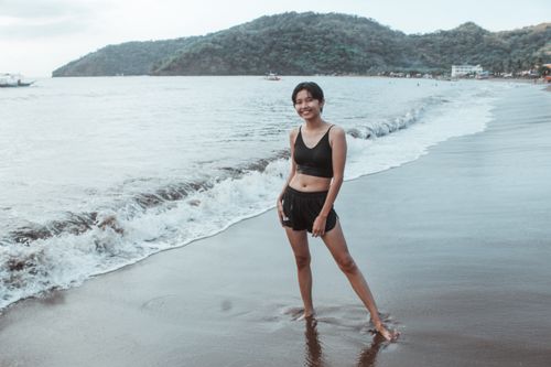 Free An Asian woman smiles while standing barefoot on a scenic beach shoreline. Stock Photo