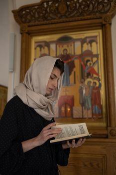 Woman in headscarf reading book in a church, standing before religious icon.