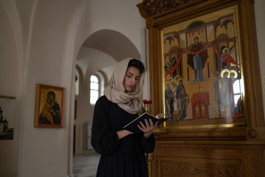 A woman in a headscarf reading a book in an Orthodox church, surrounded by religious icons.