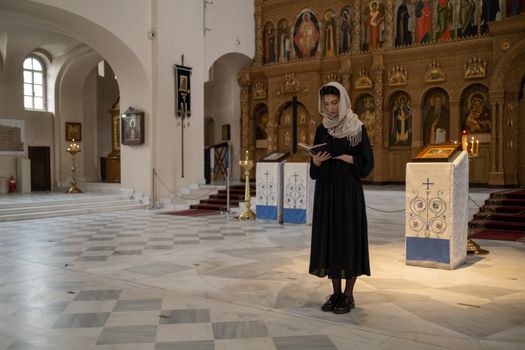 Woman in traditional attire reading book in an ornately decorated Orthodox church.