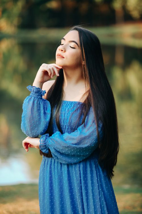 Free Portrait of a contemplative young woman in a blue dress, enjoying nature by a serene lake. Stock Photo