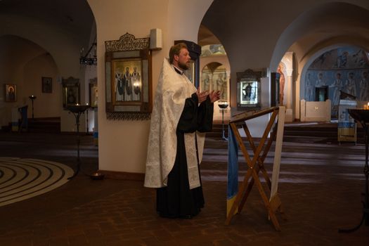 Orthodox priest in vestments standing and praying in an ornate church interior.