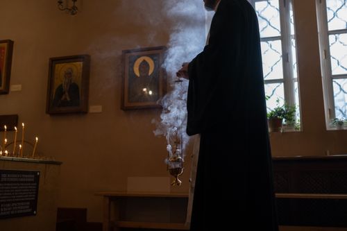 Free Priest holding censer with incense in a dimly lit church, surrounded by religious icons and candles. Stock Photo