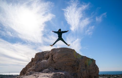 Free A person in a jacket jumps off a rocky cliff under a bright blue sky, showcasing adventure and freedom. Stock Photo