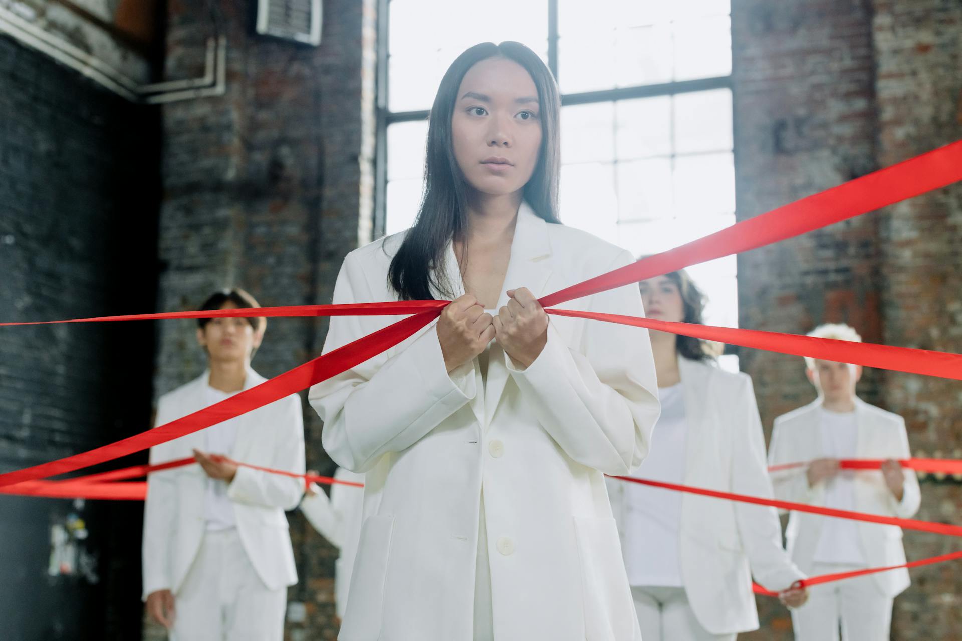 A group of adults in white suits holding red ribbons in a conceptual indoor art setting.