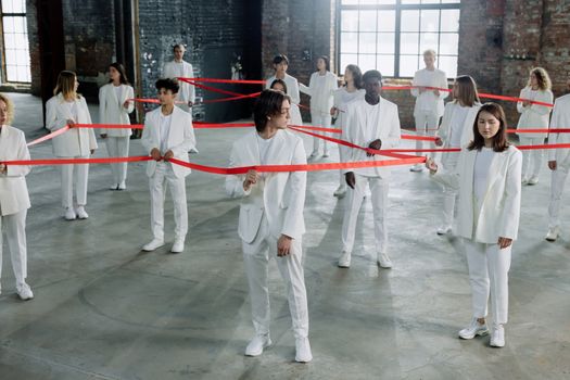 A diverse group of individuals in white clothing connected by a red ribbon indoors, symbolizing unity and diversity.