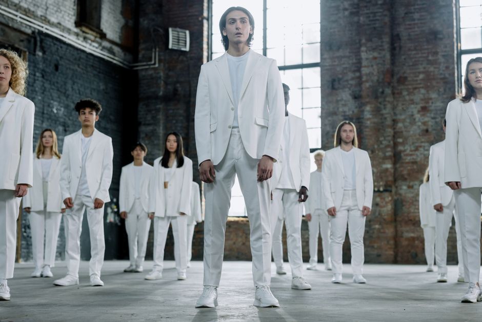 A diverse group of models in elegant white suits standing in an industrial indoor setting.