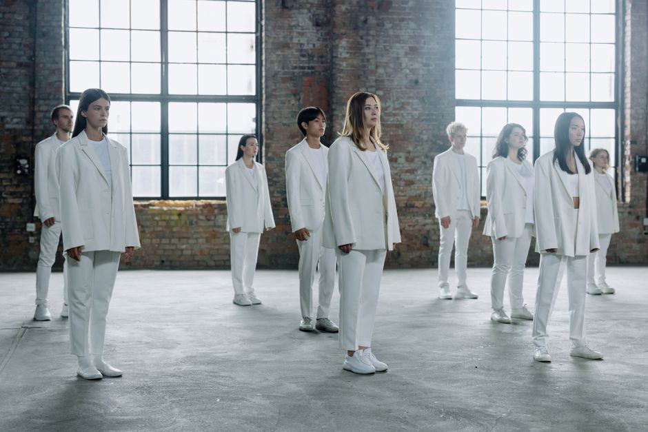 A group of diverse individuals in white outfits standing seriously in a brick-walled warehouse.