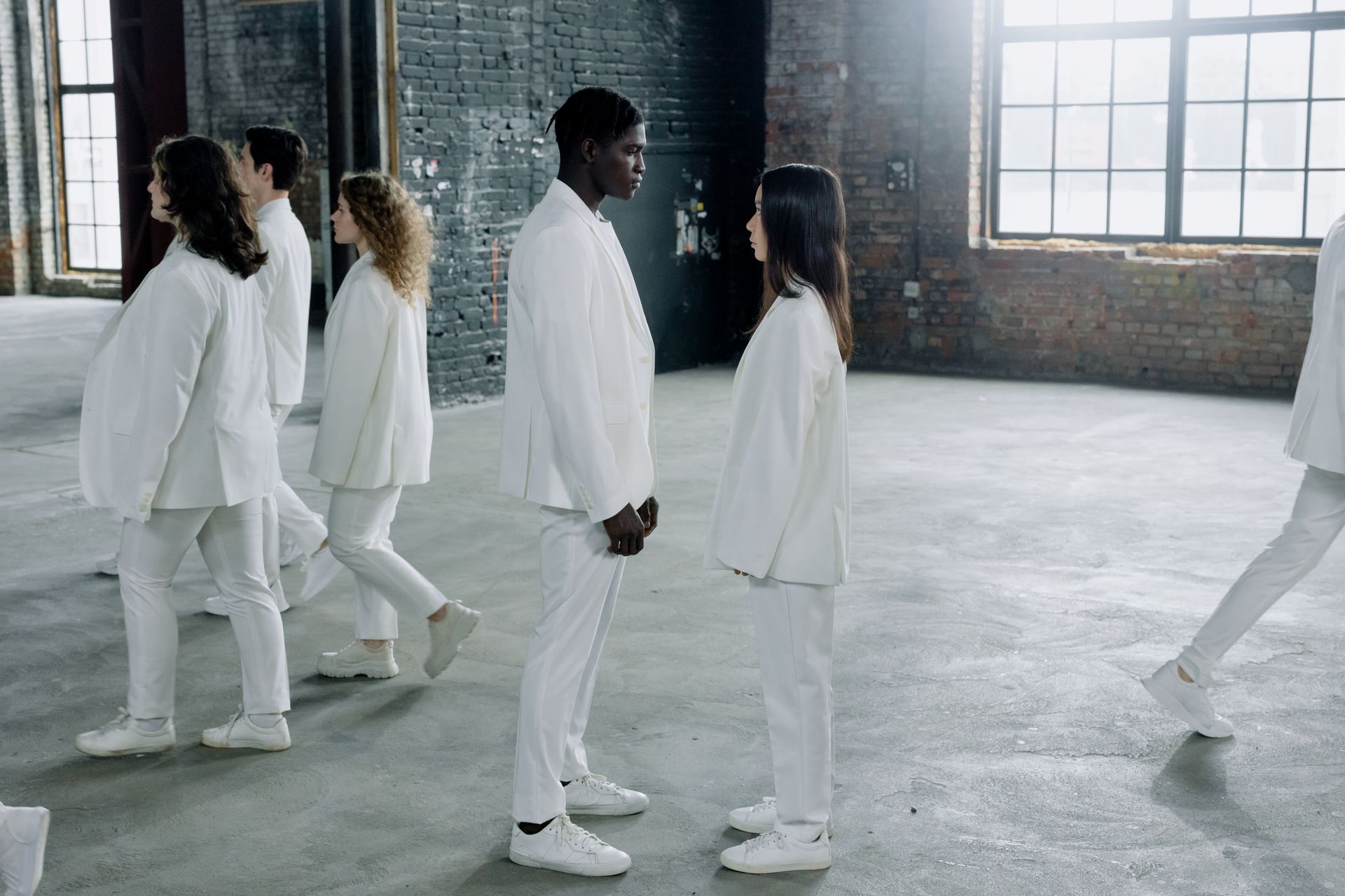 A diverse group of adults in white suits stands and walks in an industrial space with brick walls.