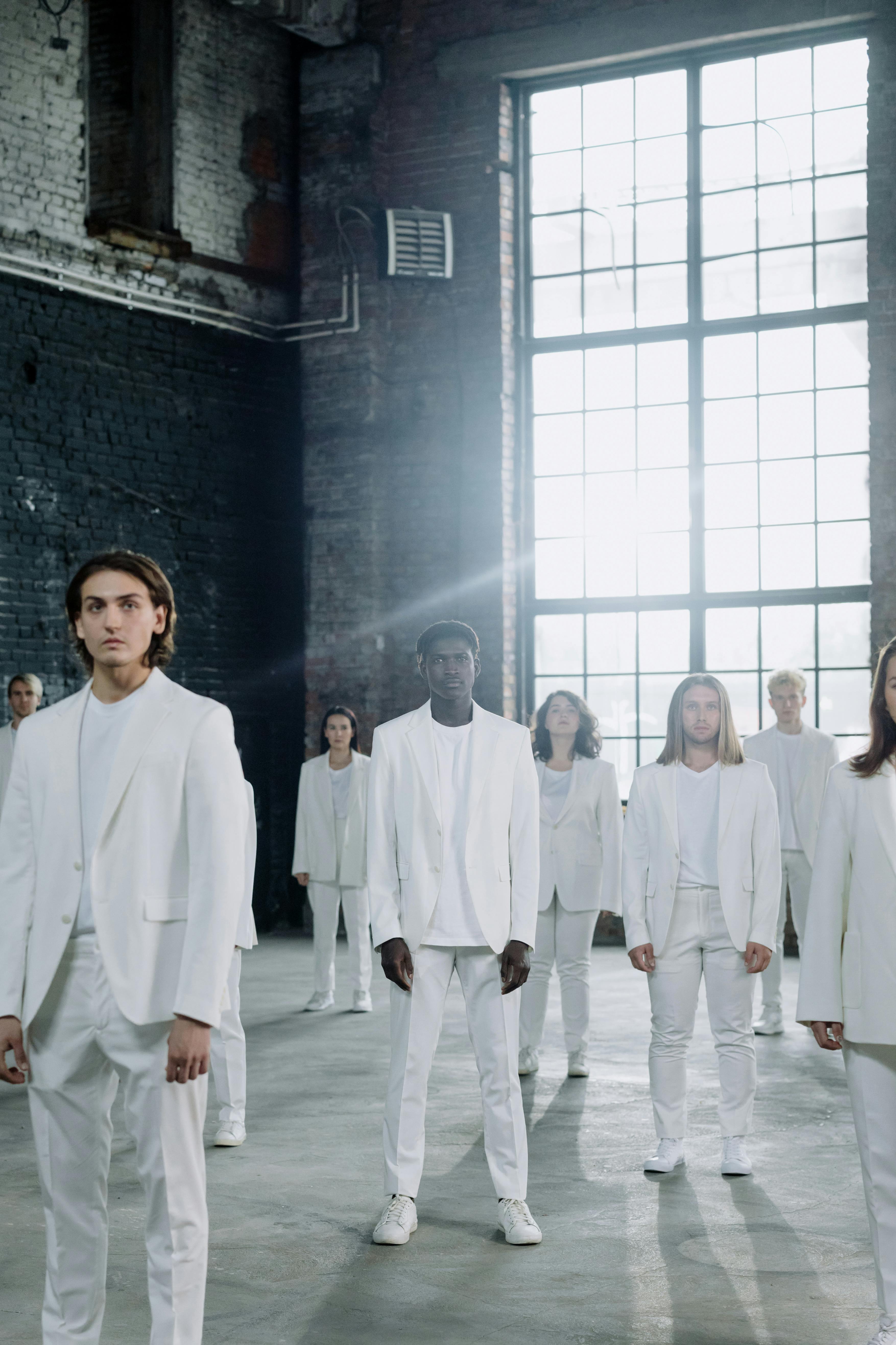 Group of diverse models in white suits posing in industrial space with sunlight.