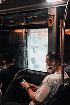 Passengers wearing masks using phones on a rainy night bus in Istanbul.