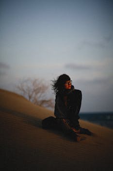 Woman with glasses sitting on a sand dune at twilight, capturing a serene and contemplative moment.