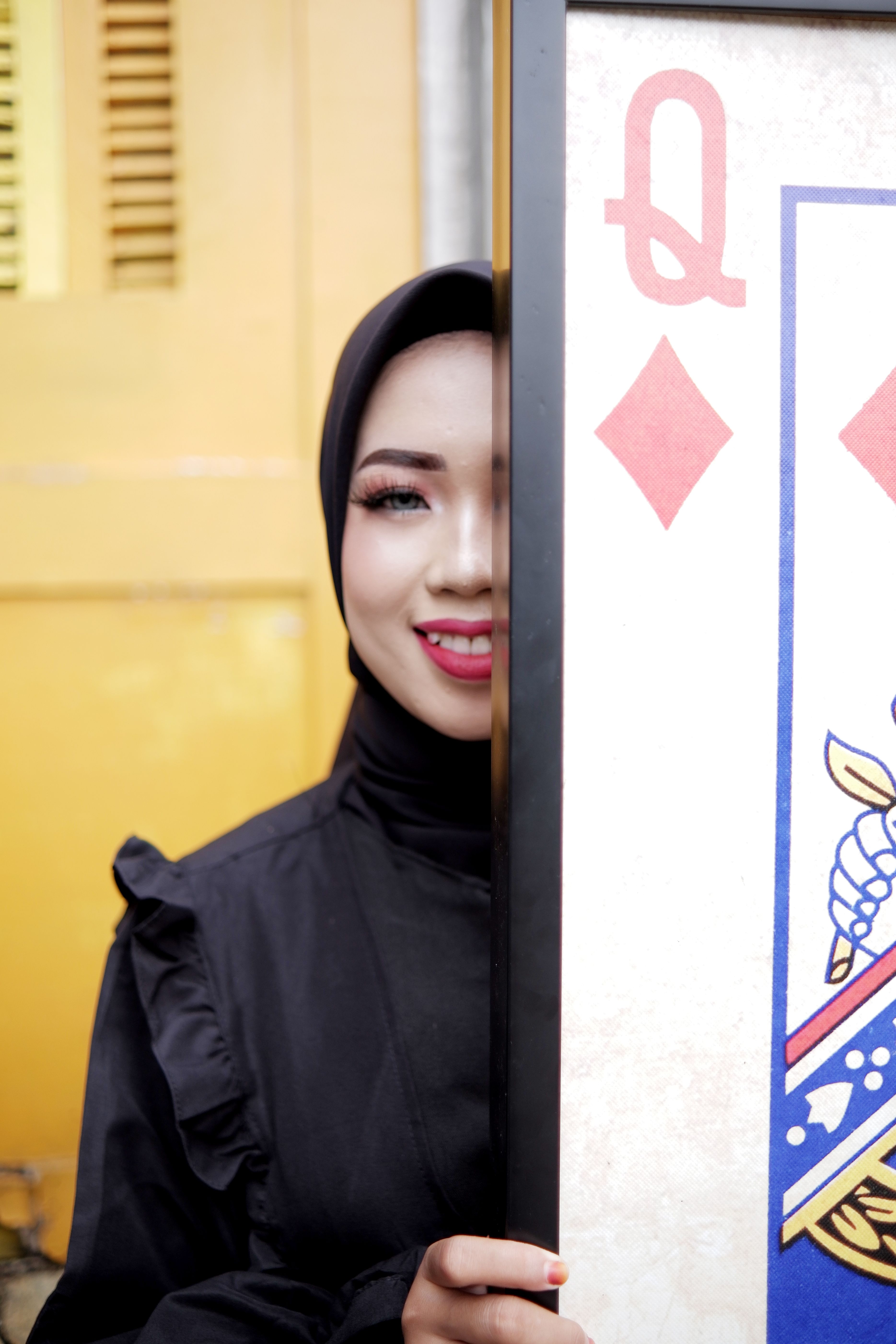 A fashionable woman in a hijab smiles beside a large Queen of Diamonds playing card.