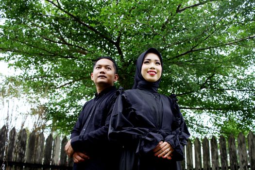 A couple in traditional clothing stands together outdoors under a tree.
