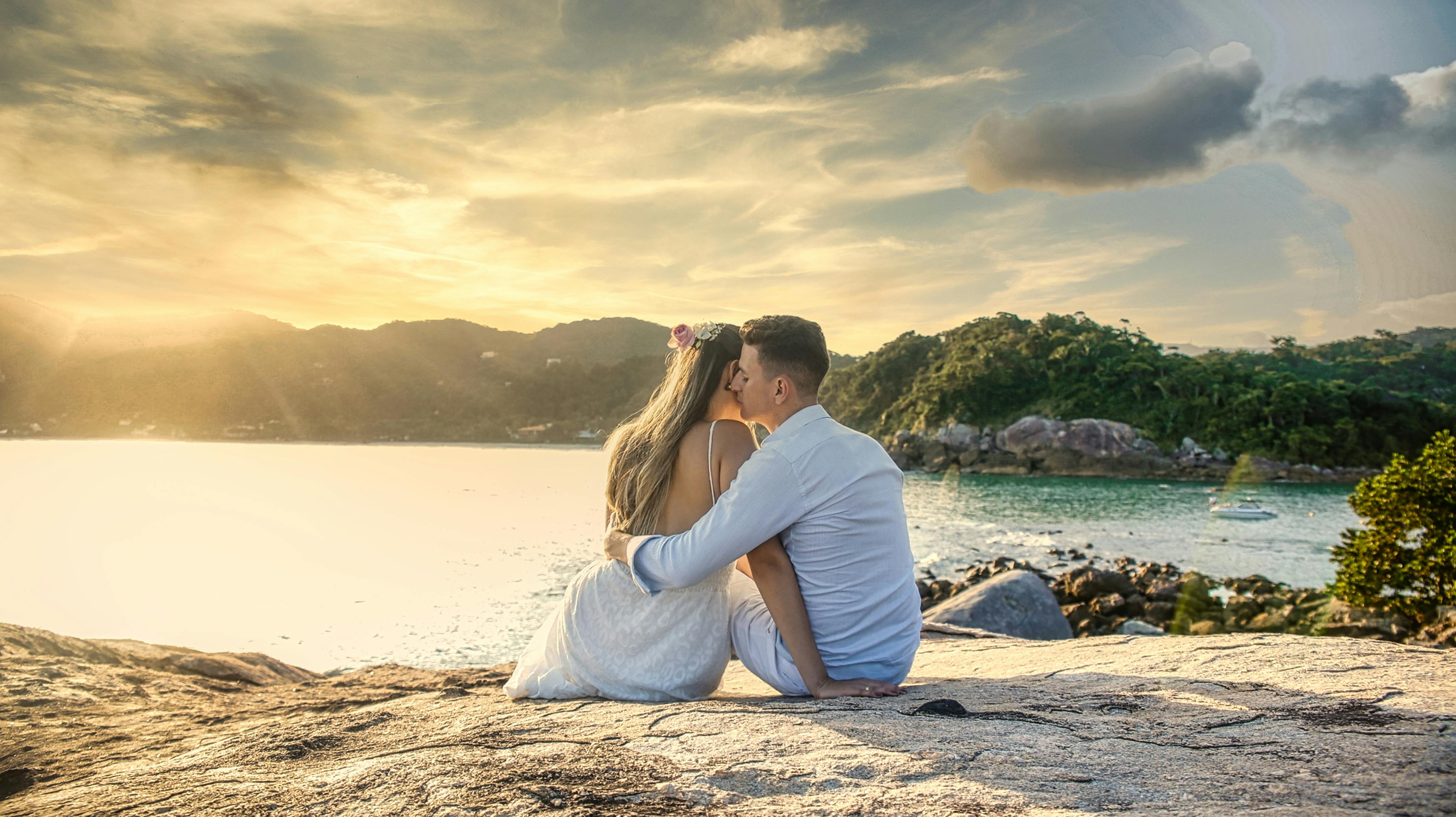 couple in white clothing sitting on rock near water