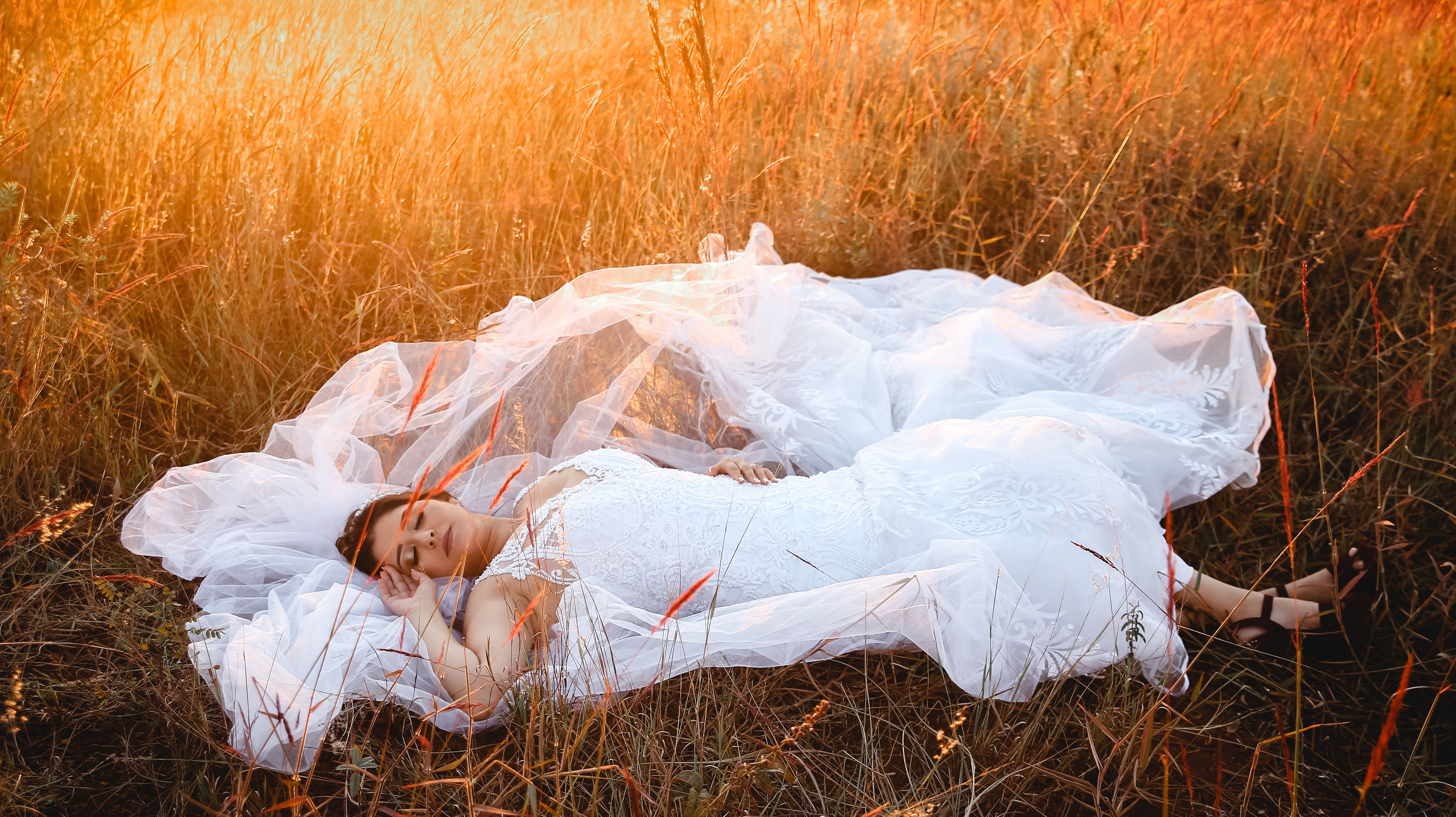 Serene bride in elegant gown resting amidst a sunlit field, capturing a peaceful moment.