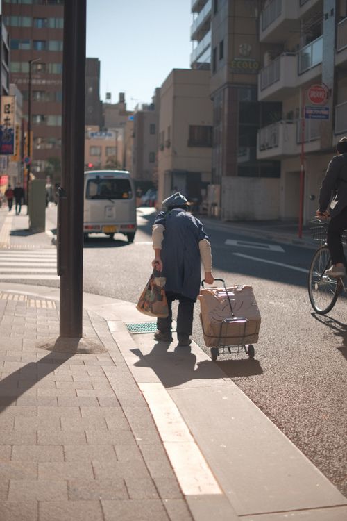 Free An elderly person pulls a trolley down a sunlit city street, capturing daily urban life. Stock Photo