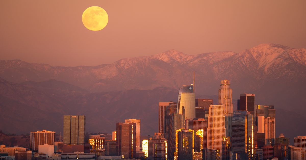 Stunning view of the Los Angeles skyline with a full moon rising over skyscrapers at sunset.