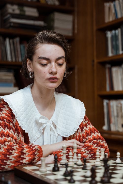 Free A young woman focused on playing chess in a library setting, showcasing concentration and strategy. Stock Photo