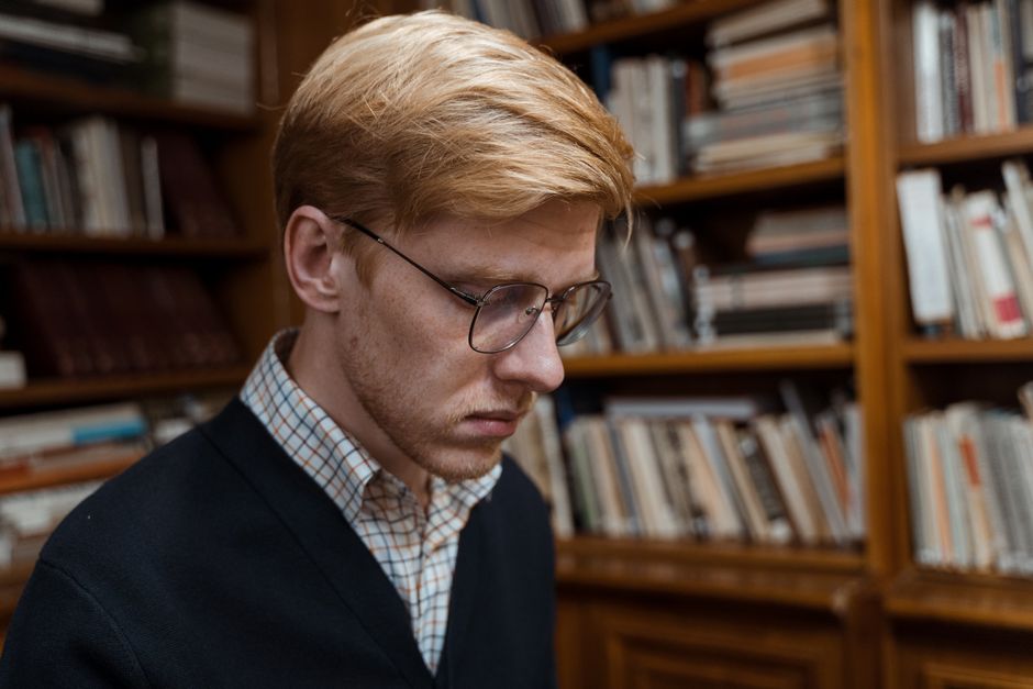 A young man with glasses focused on reading in a library environment, surrounded by books. A young man with glasses focused on reading in a library environment, surrounded by books.
