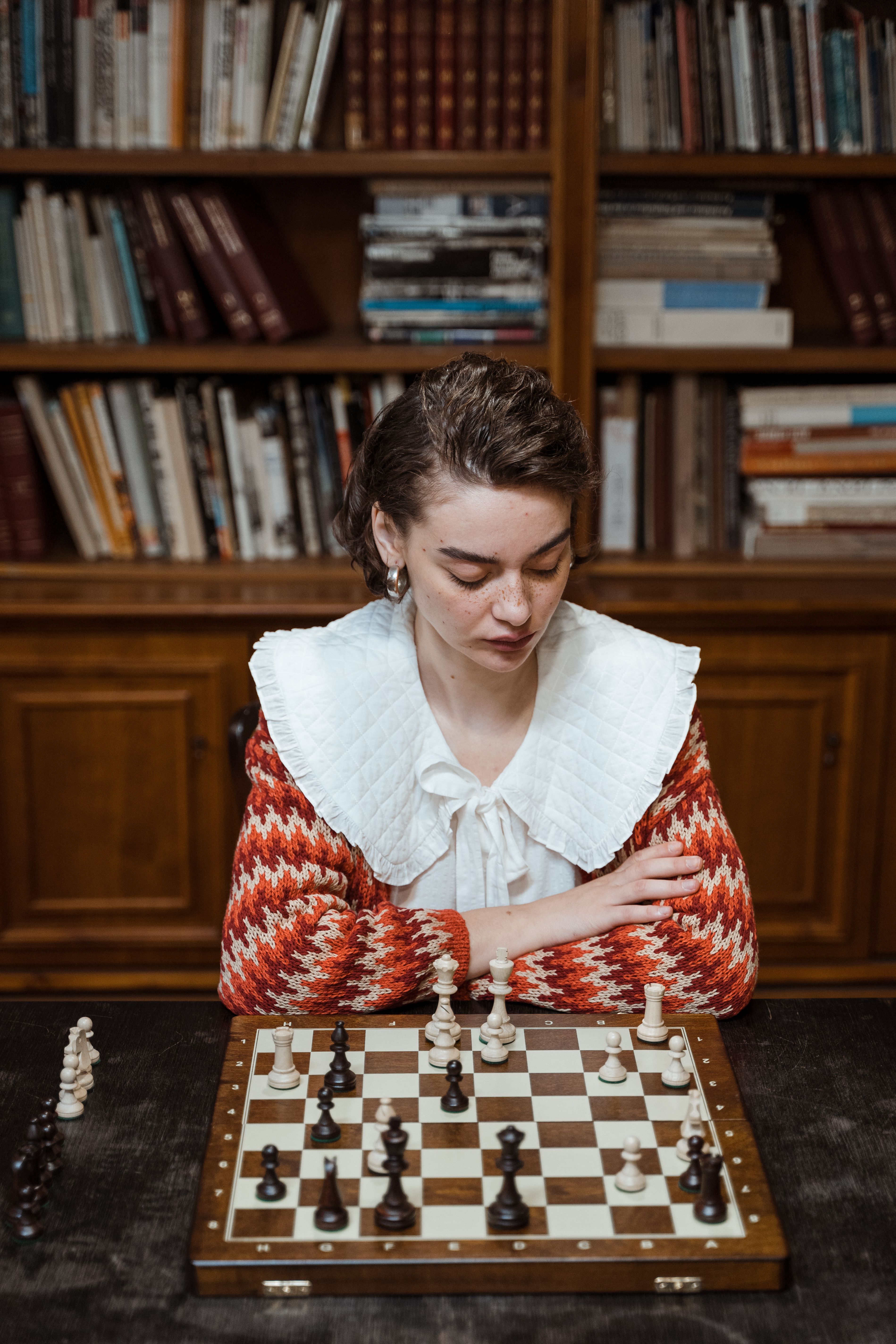 Focused woman plays chess in a cozy library setting, pondering her next strategic move.