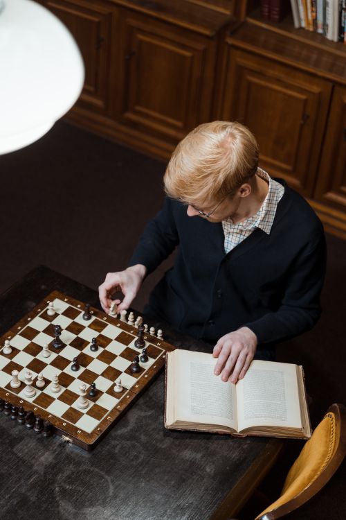 Free A man engrossed in a chess game with an open book nearby on a wooden table. Stock Photo
