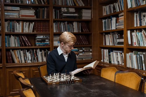 Free A man reads a book at a library table with a chessboard, surrounded by shelves of books. Stock Photo