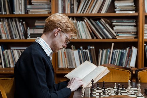 Free Side view of man reading and playing chess in a library setting. Stock Photo