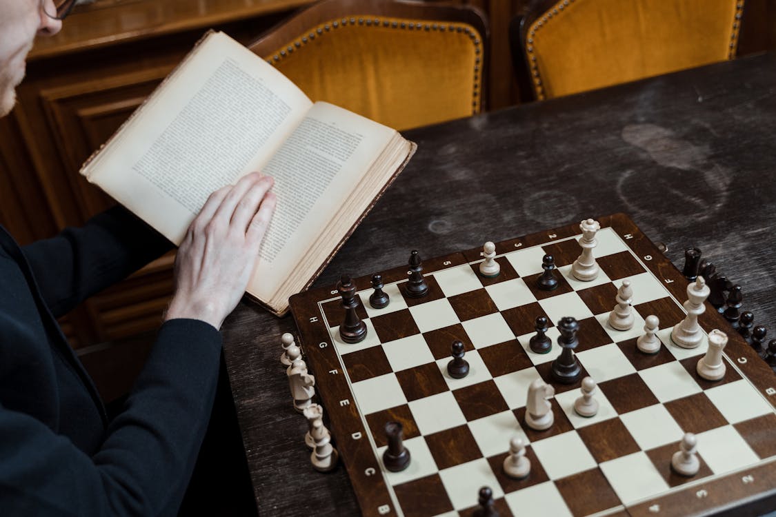 Person Reading Book on Table beside Chess Game 