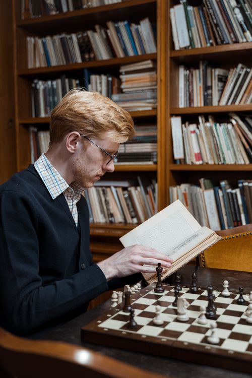 Free A man reading a book beside a chessboard in a cozy library setting. Stock Photo