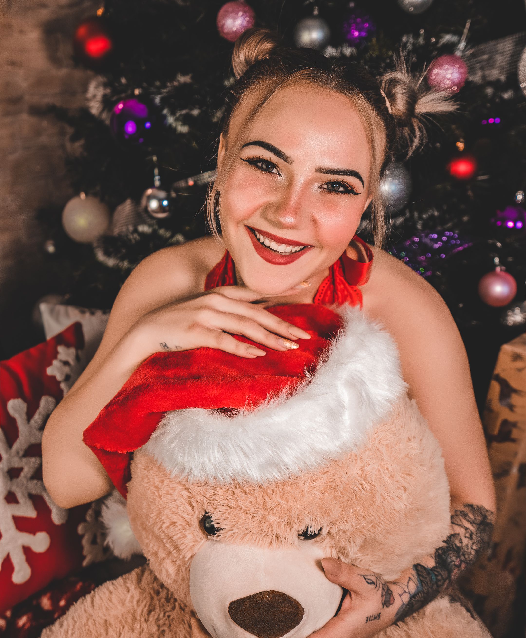 Joyful woman in festive attire hugging a teddy bear by a decorated Christmas tree.