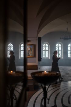 A parishioner walks through a serene, dimly-lit Orthodox church hallway, reflecting in a mirror.