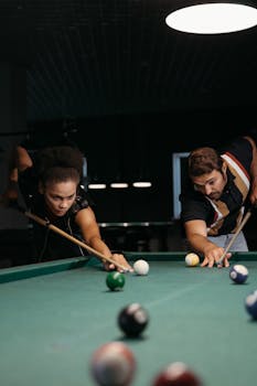 A focused match between two adults playing billiards in a dimly lit room using cue sticks.