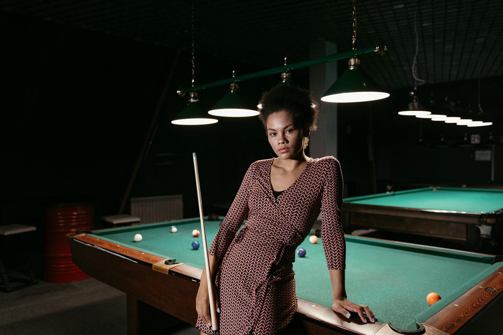 Stylish woman with afro hair posing confidently by a pool table in a dimly lit billiard room.