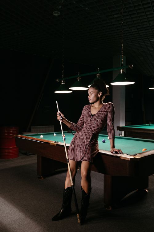 Free A fashionable woman in boots poses confidently by a billiard table in a dimly lit room. Stock Photo