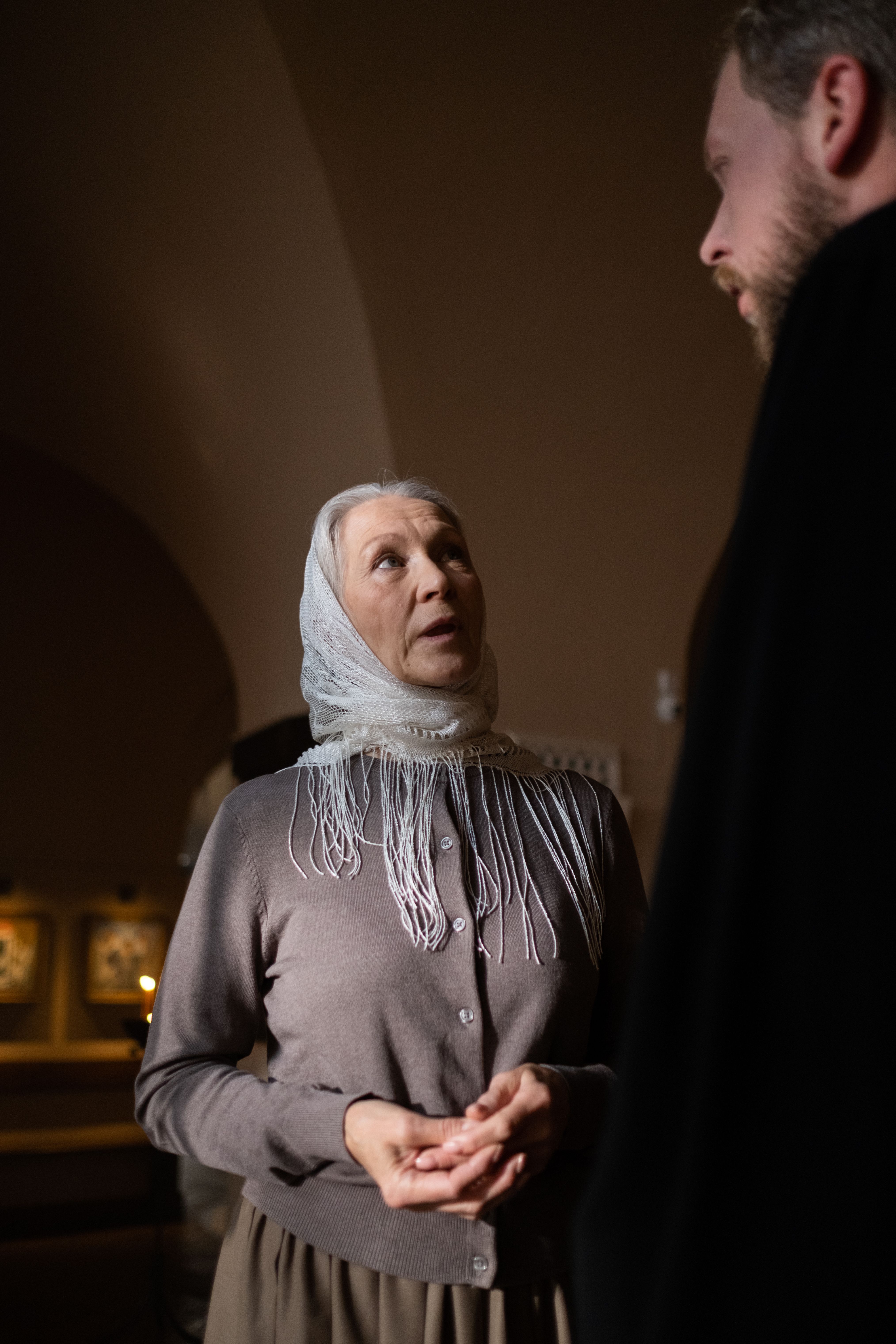 An elderly woman wearing a headscarf converses with a bearded man in a dimly lit room.